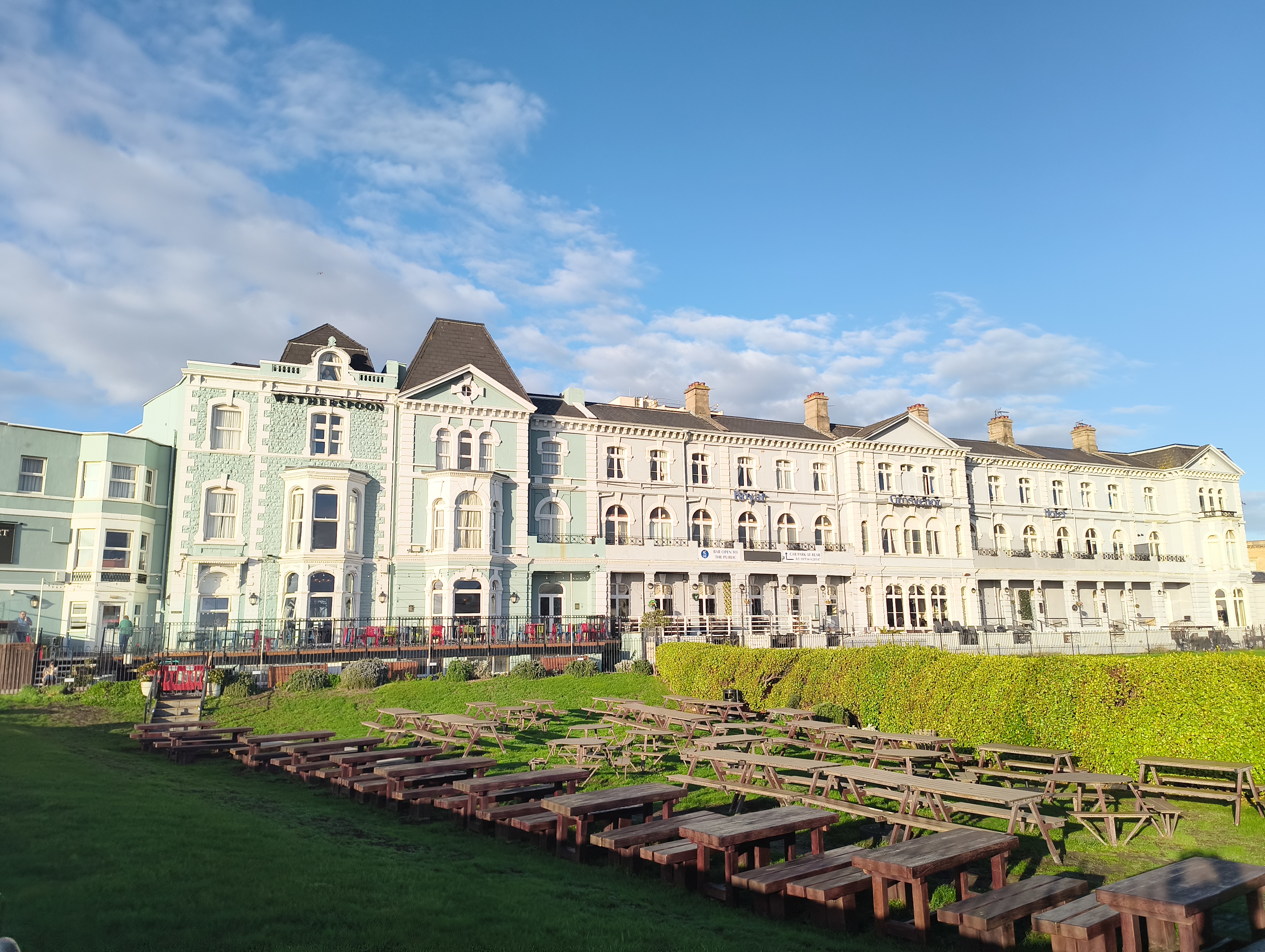 A long, historic hotel with a distinctive mint-green and white façade, signed "Wetherspoon," overlooks a tiered, grassy area filled with rows of empty wooden picnic tables.