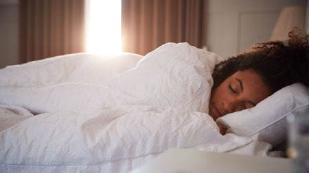 A woman sleeping soundly in bed under a white duvet, as early morning sun shines through the curtains behind her.