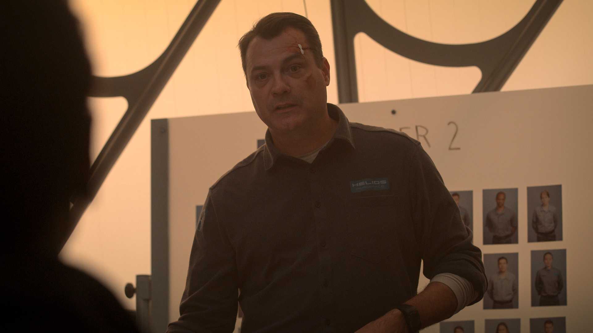 A man who has been roughed up (bruise on cheek, stitches on eyebrow) is standing in front of a whiteboard with several images of other crew members.