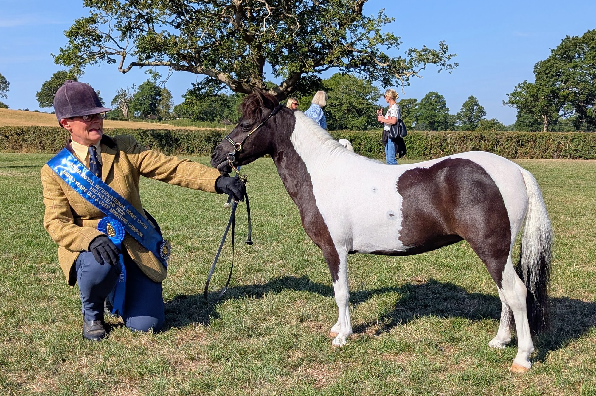 A piebald Miniature Horse with his handler, who is crouching down wearing tweed with her blue winner&amp;rsquo;s sash and rosette
