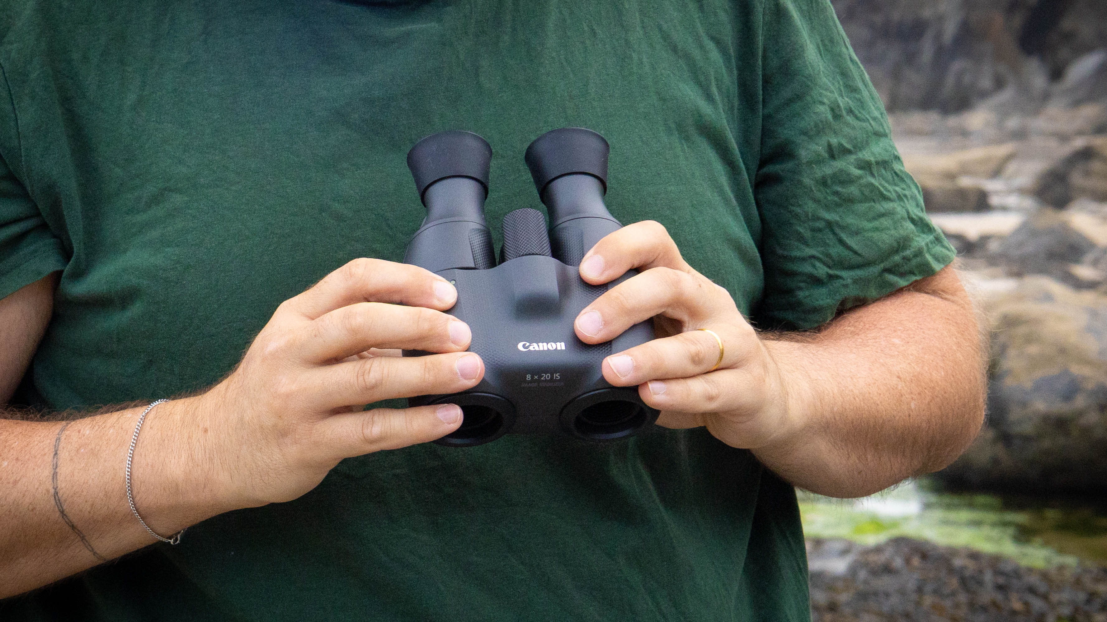 A male holding the Canon 8x20 IS binoculars close to his chest, showing the Canon logo on the binos.