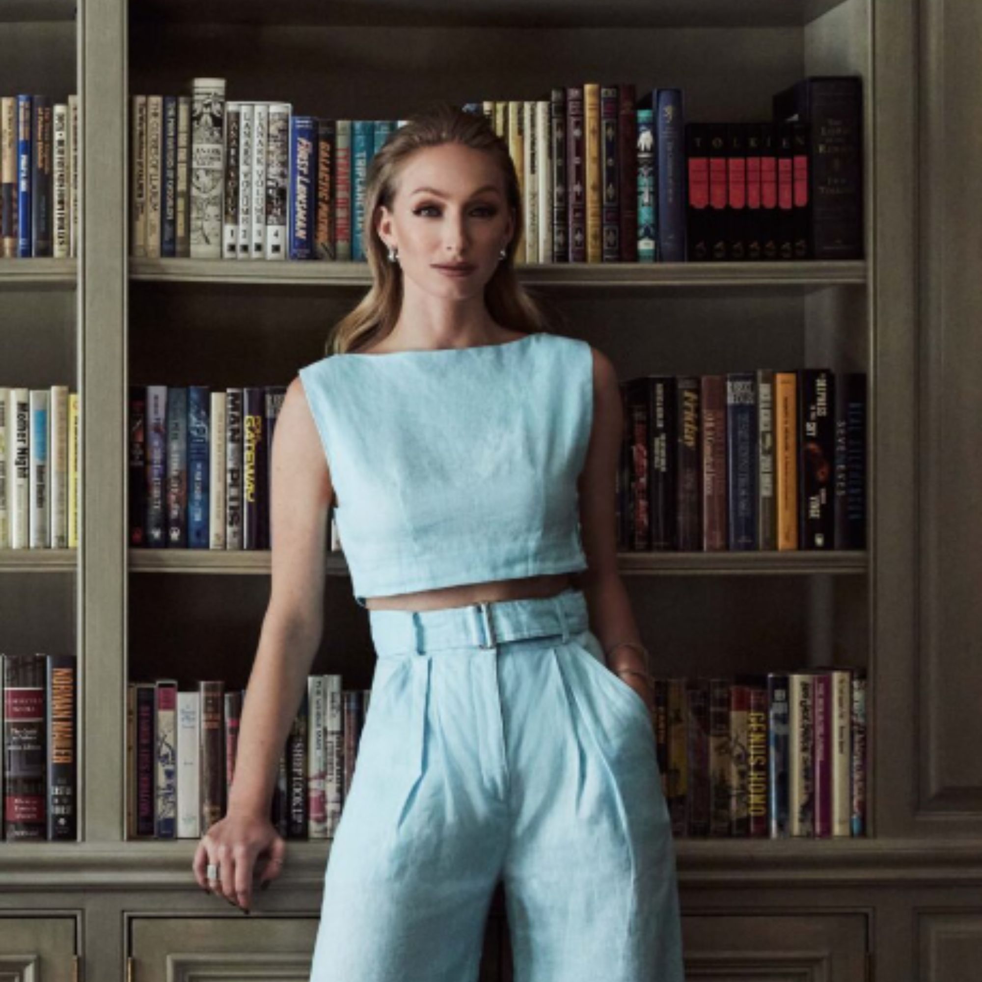 a blonde woman standing in front of a green bookcase in a matching light blue set