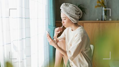 Woman wearing towel on her head after getting out of the shower, brushing teeth and looking at phone in bedroom to find out should I brush my teeth before breakfast