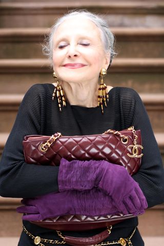 A photo of Joyce Carpati in an over 60 outfit featuring a black long-sleeve top, purple gloves, a quilted Chanel bag, a gold belt, and gold earrings.