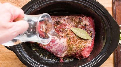 A raw cut of beef brisket in a slow cooker, flavored with herbs and spices, and a hand pouring liquid