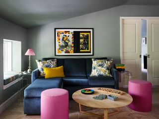 Image of a muted moss green living room with a navy blue, velvet small sectional sofa. There are two small, round ottomans in a hot pink shade and a small pink lamp on the side table next to the sofa. There is a large round wooden coffee table with games on it.