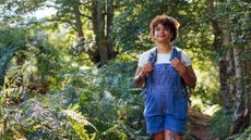 woman wearing denim short dungarees and a white tshirt walking in a forest setting.