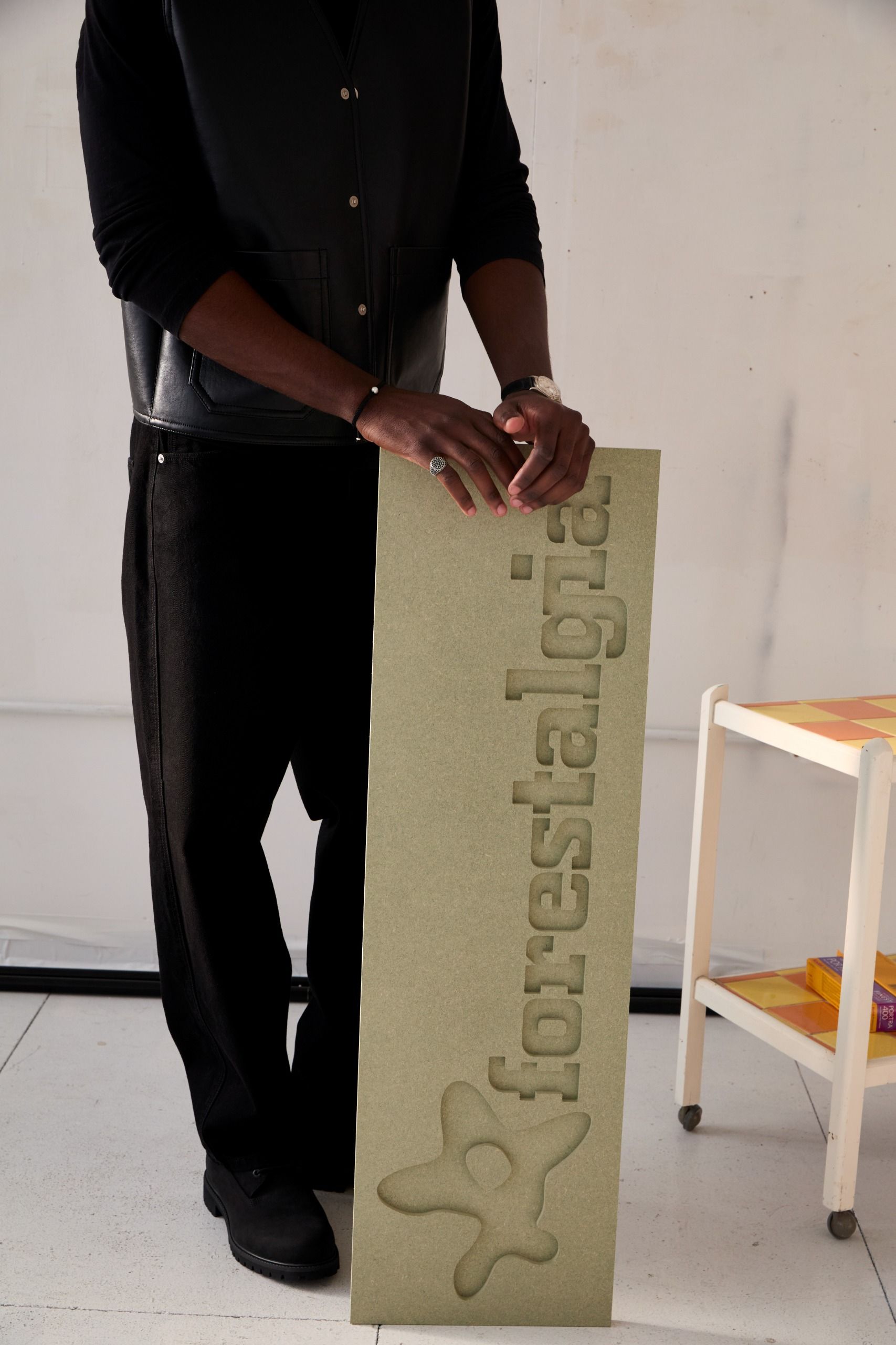 A young man poses in his industrial studio dressed in casual smartwear, sitting or standing in front of a gray wall with his sculptural creations.