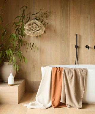 bathroom with large pale wooden plans on floors and walls, freestanding bath, hanging wicker pendant light, black wall mounted shower and taps