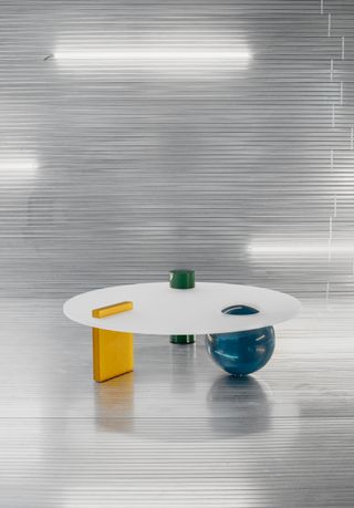 The round deep green, rectangular orange, and ball-shaped blue volumes are supporting the white circular tabletop and emerge on top of it.