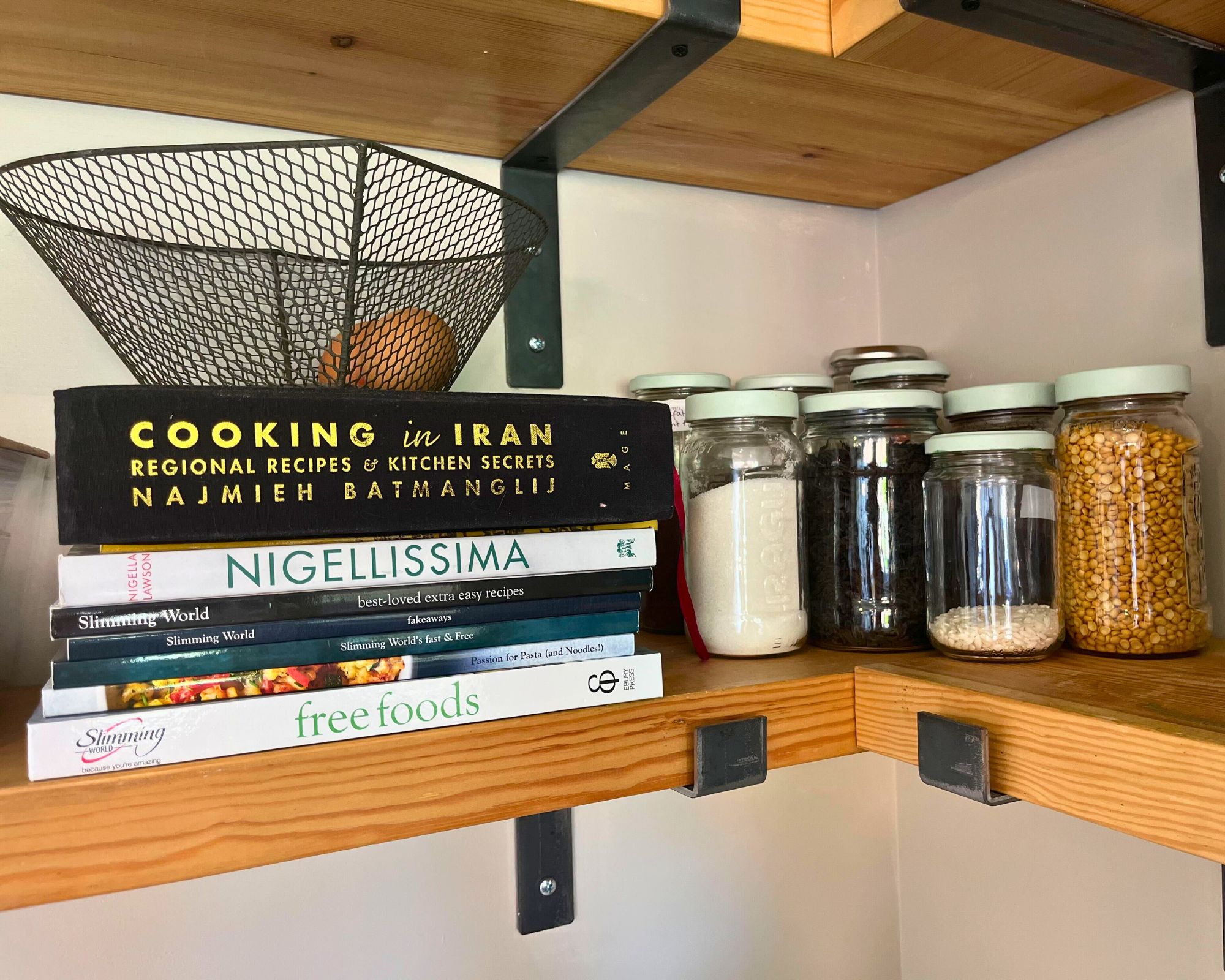 Repurposed glass jars with matching painted lids on a wooden pantry shelf with industrial black metal brackets and a pile of cookbooks, including Cooking in Iran and Nigellissima
