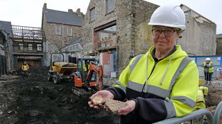 An excavator holds up two tiles discovered at the site that likely holds the remains of Friary of St. Saviour, a medieval friary in Pembrokeshire, Wales. Behind her you can see three old brick houses and a road that's been completely dug up by a mini digger.