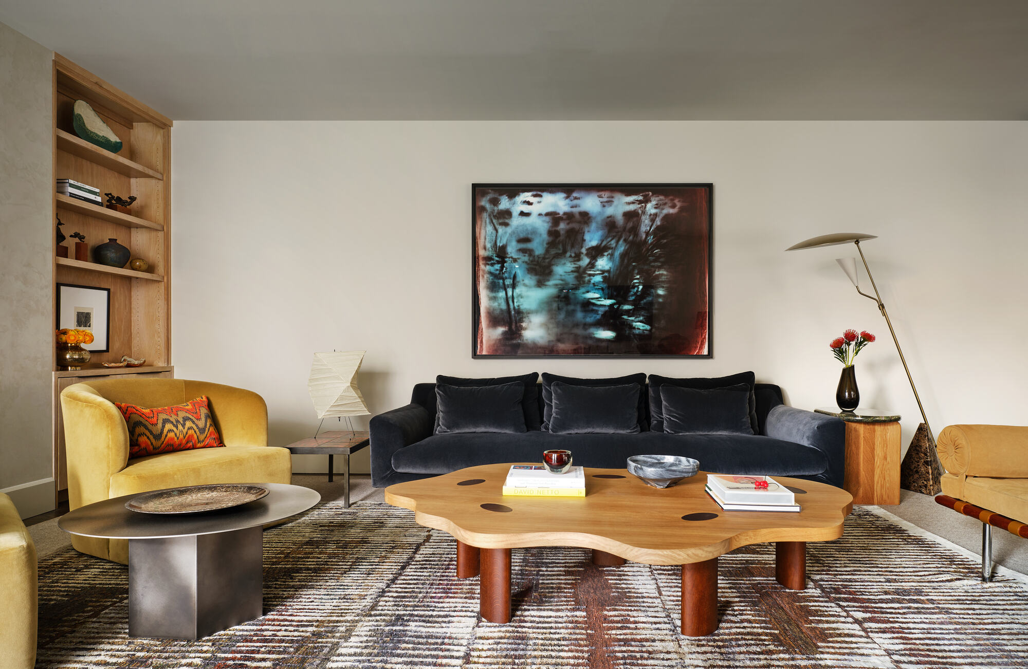 modern living room with gray velvet sofa, artwork, yellow velvet armchair, chrome side table, timber wiggly coffee table, accent lighting, patterned rug and built-in bookshelves