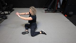 Woman demonstrates 90/90 exercise. She sits on the floor facing left, with both legs on the floor with her knees bent to 90°. She wears a black T-shirt, black leggings and black trainers.