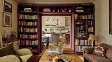 Living room with a built-in bookcase surrounding the doorway, painted burgundy, filled with books and a built-in bar. The living room features a wooden coffee table with candles, a vase, and books, a velvet green armchair, a brown sofa and looks through onto the kitchen
