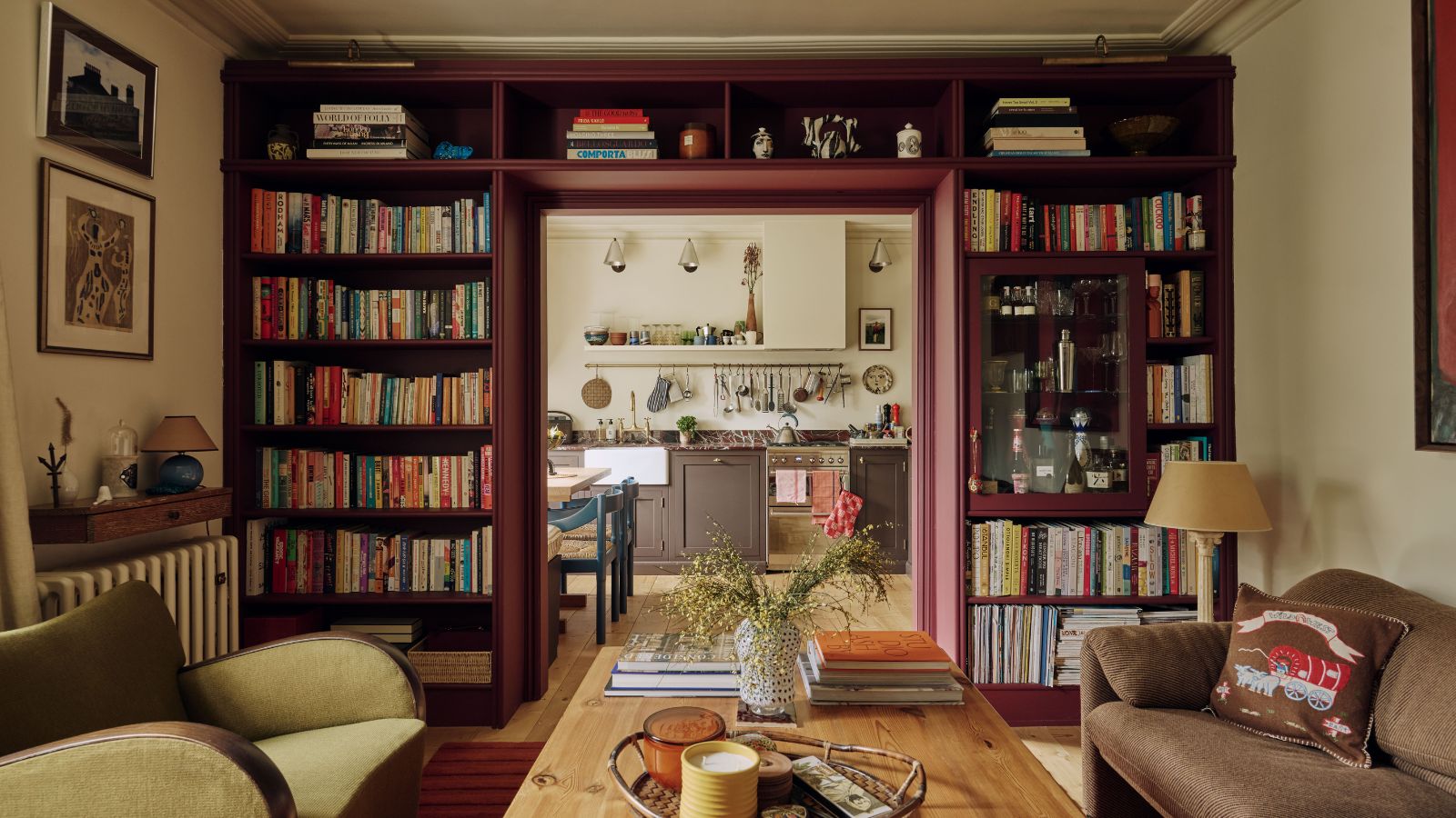 Living room with a built-in bookcase surrounding the doorway, painted burgundy, filled with books and a built-in bar. The living room features a wooden coffee table with candles, a vase, and books, a velvet green armchair, a brown sofa and looks through onto the kitchen