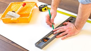 Hans measuring wallpaper and drawing a straight edge with yellow wallpaper tray and brush 