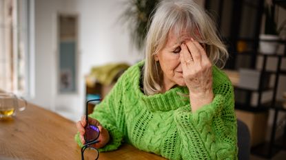 An older woman sitting at her dining room table rubs her forehead like she regrets making a mistake.