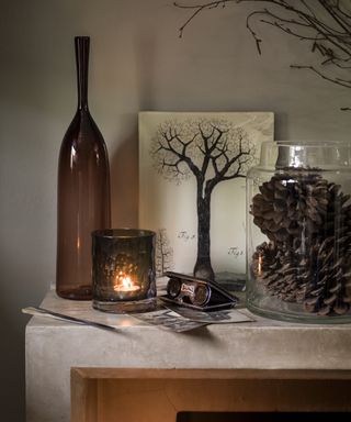 Fall mantel ideas with brpown glass bottle and candle holder, pine cones in a hurricane vase and tree painting