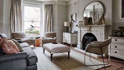 Neutral living room with large white mantelpiece, circular mirror, white rug, and grey sofa. In the centre is a striped footrest. There are large bay windows looking over a street behind in the back.