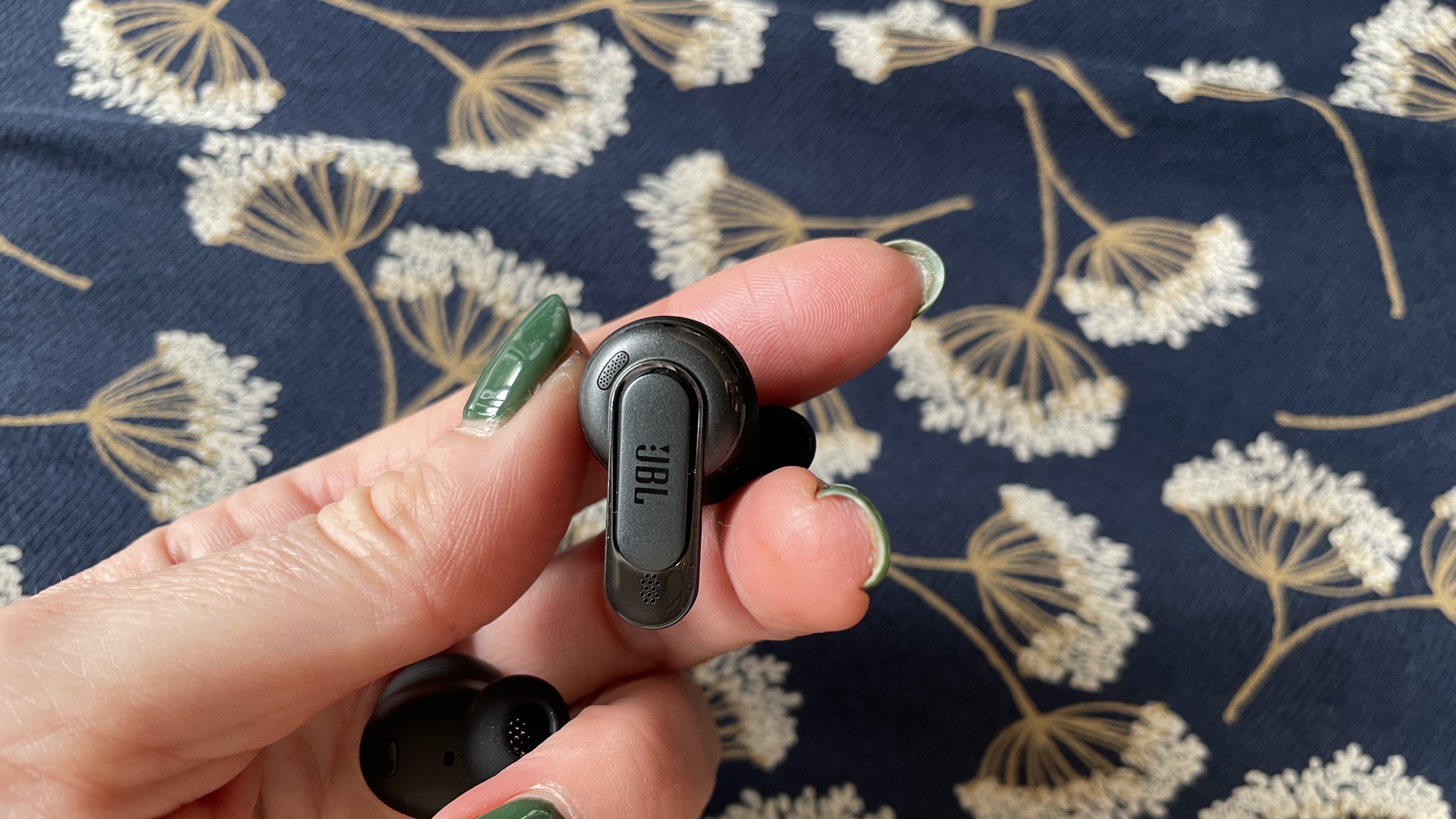 JBL Tour Pro 3 earbuds held in a hand, with navy floral background