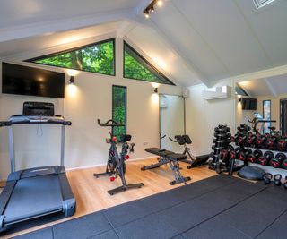 A bright garage converted into a home gym, with treadmills, bikes, and weights. Spotlights and natural light from triangular windows illuminate the room.