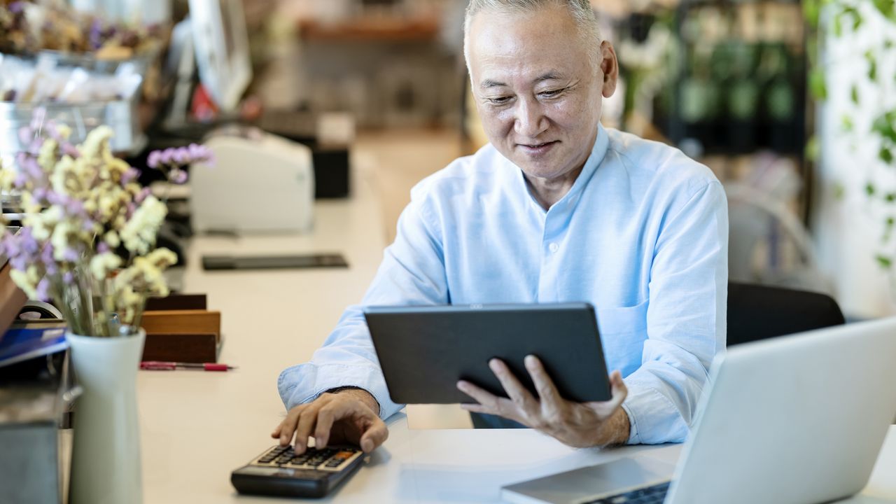 A small-business owner does some calculations on a calculator while looking at his tablet in his flower shop.