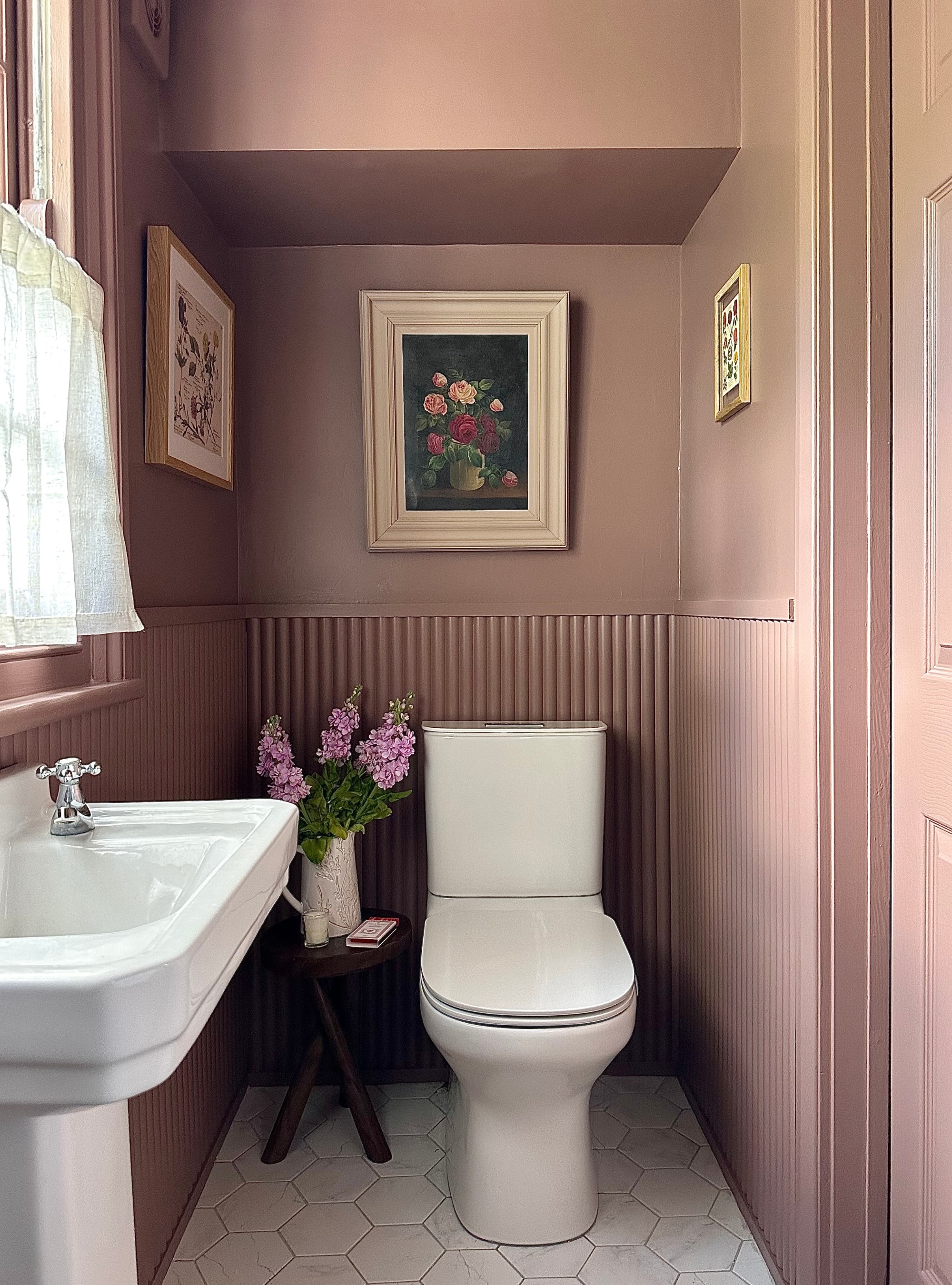 pink color drenched bathroom with botanical art above toilet, fluted wall panelling, white freestanding sink, cafe curtains, and stool with vase of flowers and magazines