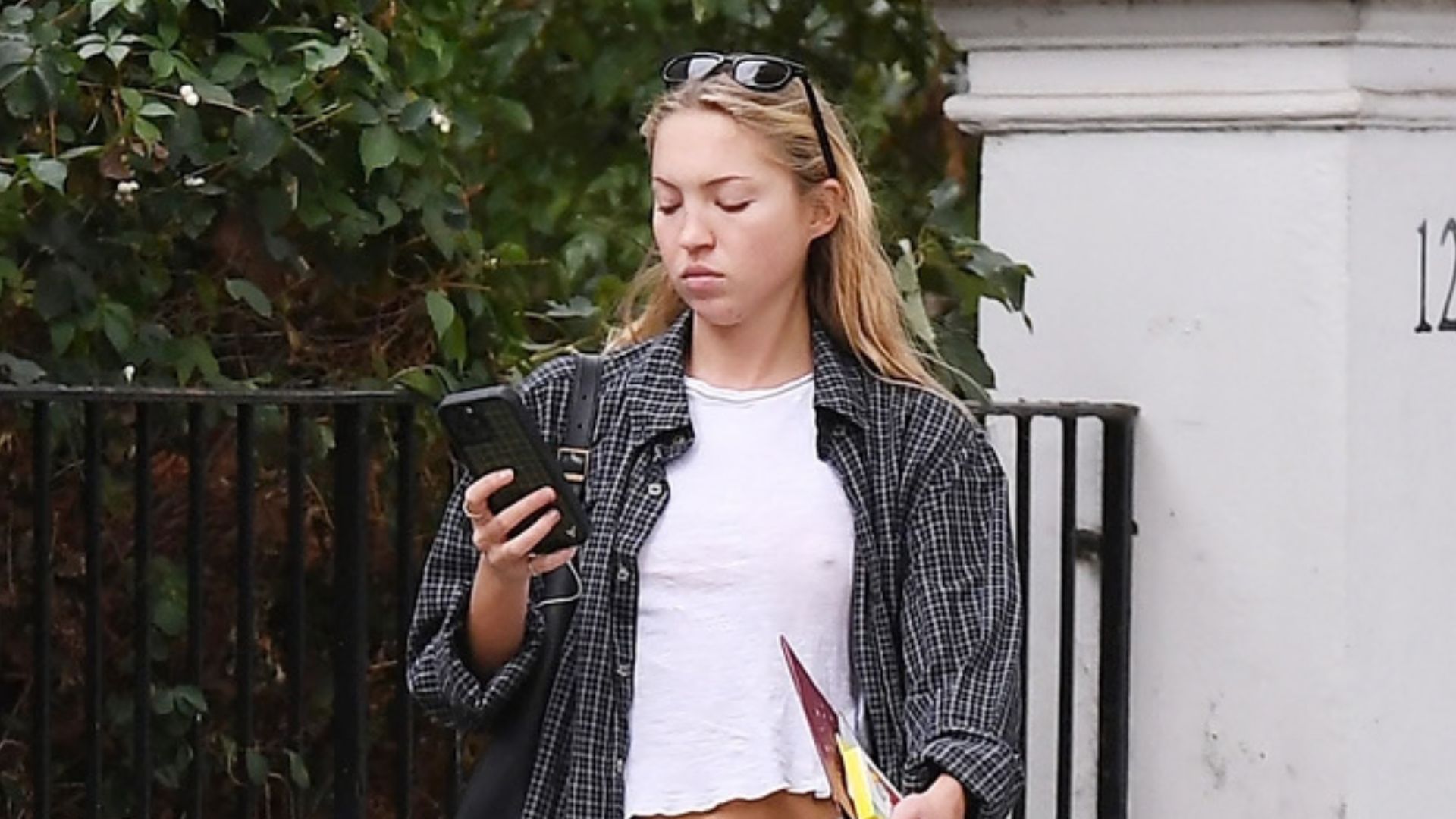 Lila Moss steps outside wearing mid-rise bootcut jeans with black ballet flats, a white tee and a flannel shirt.