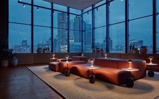 Night falls on a glassy, industrial hotel lobby, with plush leather modular sofas and mushroom-shaped, warmly lit table lamps lighting up in a beautiful, eye-catching effect.