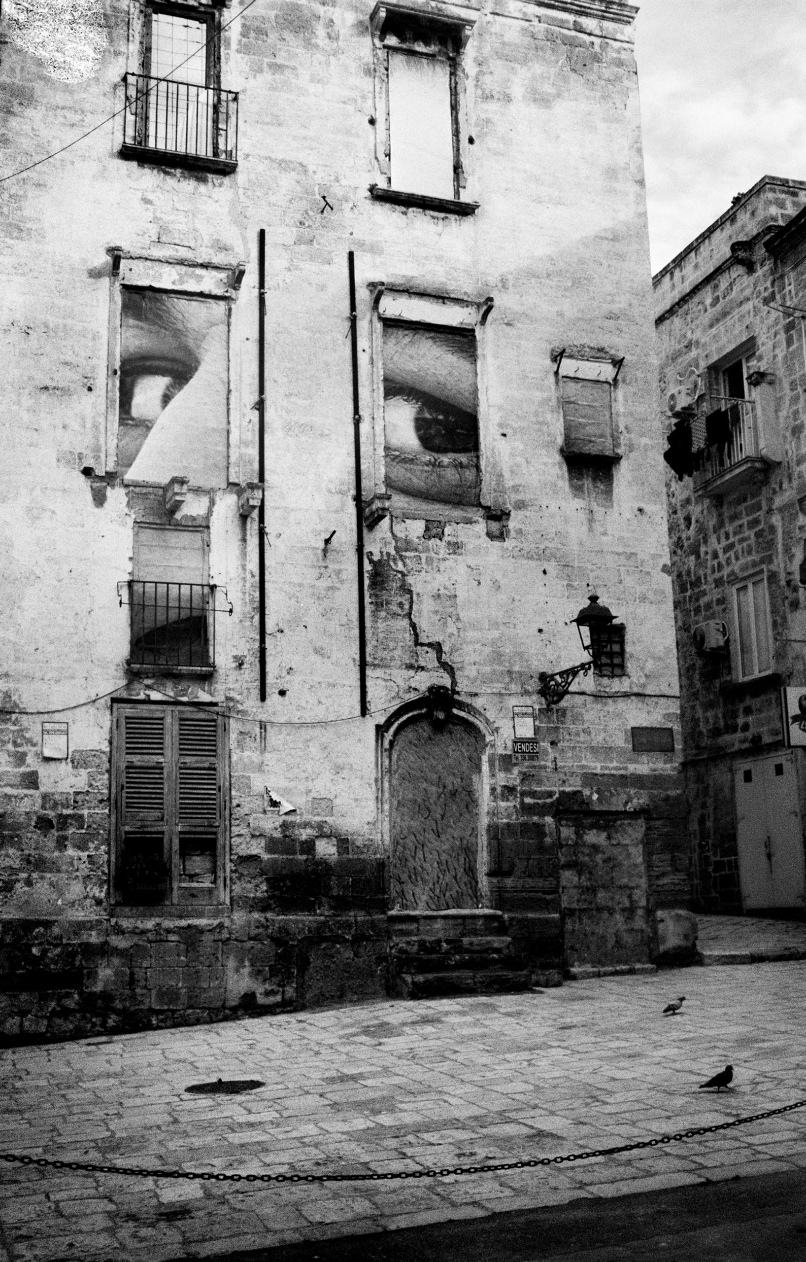 A crumbling stone building has giant photographs of eyes pasted into its empty window frames, overlooking a deserted cobbled square where two pigeons stand.
