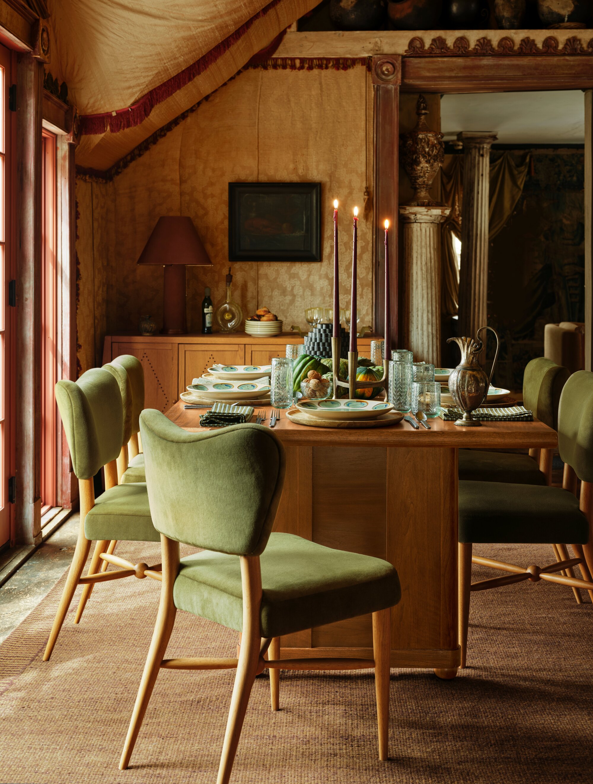 a timber dining table with green velvet chairs, a orange-toned rug, and table set with tall taper candles, green ceramic plates, napkins, and a carafe