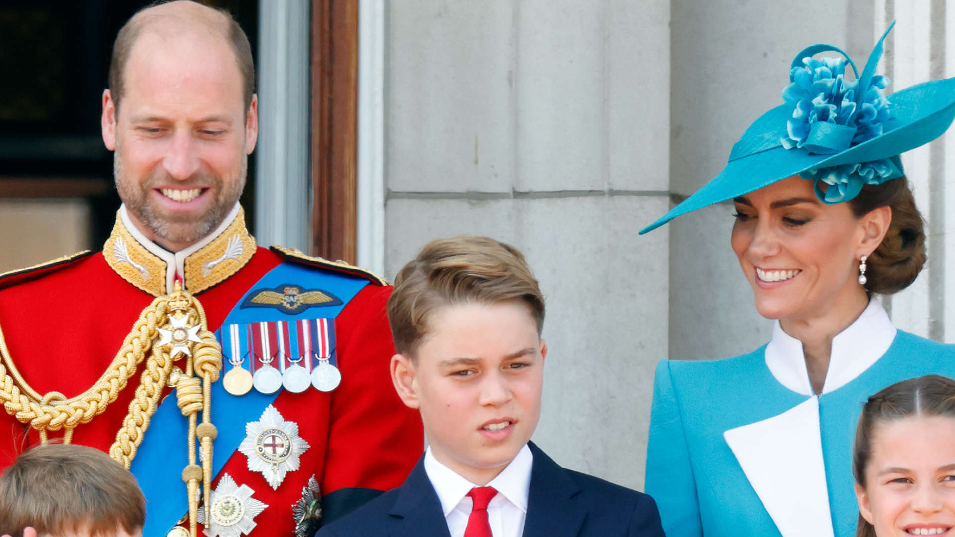 Prince George wears a navy suit with a red tie and stands on the balcony with his family