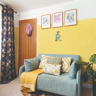 A living room painted in yellow with a small two-seater sofa and floral eyelet curtains on the window