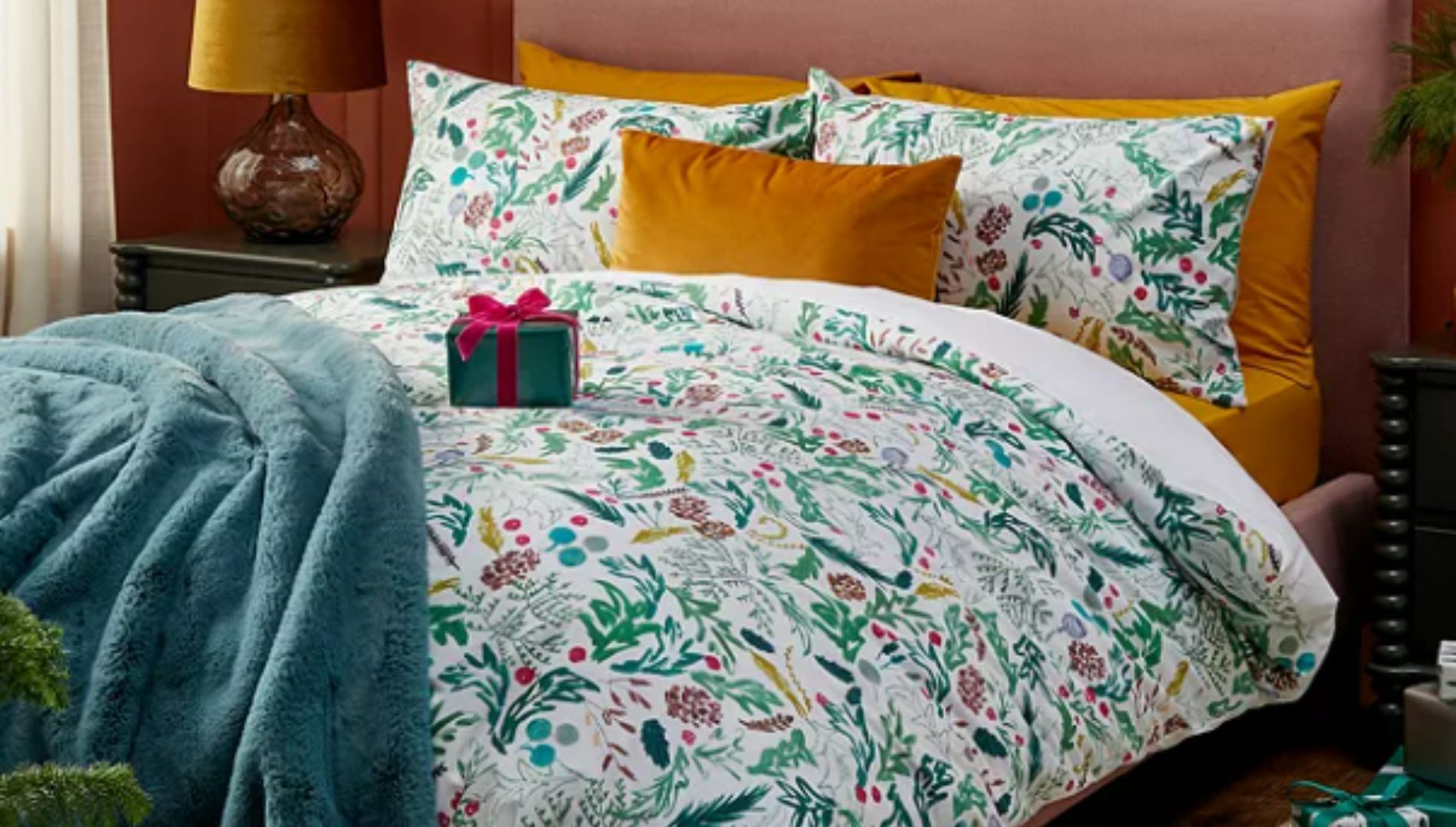 A green-and-white bedding set from John Lewis with a foliage pattern, on a double bed with orange pillows, a wrapped present and a green throw.