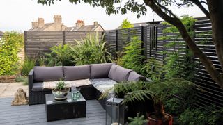 A modern grey corner sofa in a shady decked area of a garden next to a horizontal slatted fence in a dark grey shade