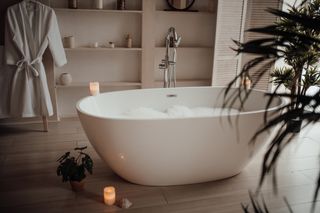 A luxury bathroom with a free standing white bath filled with bubbles and surrounded by a few plants and candles.