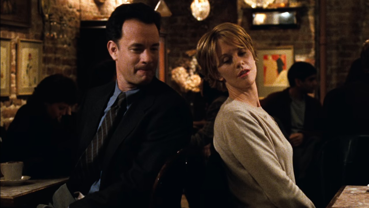 डावीकडून उजवीकडे: टॉम हँक्स खुर्चीवर बसला आहे आणि मेग रायनशी बोलण्यासाठी मागे फिरत आहे जो त्याच्यापासून दूर आहे.