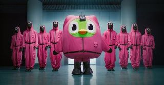 The Duolingo bird wearing the iconic pink Squid Game jumpsuit, posing with other masked guards