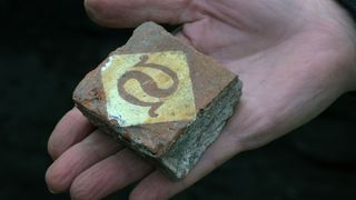 A close-up photo of a floor tile found during the excavation in Pembrokeshire, Wales. The orange-brown tile is square in shape and fits into the palm of a human hand. On the tile is a white diamond, and inside this there is what looks like to be a backwards S shape.