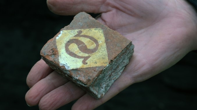Close-up photo of a floor tile found during excavations in Pembrokeshire, Wales. The square-shaped orange-brown tile fits the palm of a person's hand. On the brick is a white diamond, and inside this stone there is what looks like an inverted S shape.
