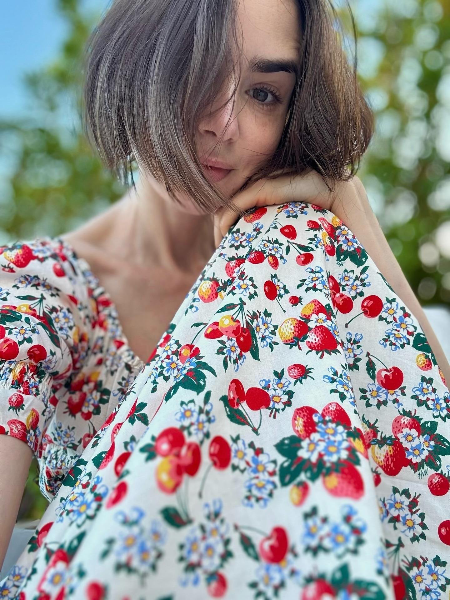 Lily Collins taking a selfie outside wearing a summer dress with a serrated bob