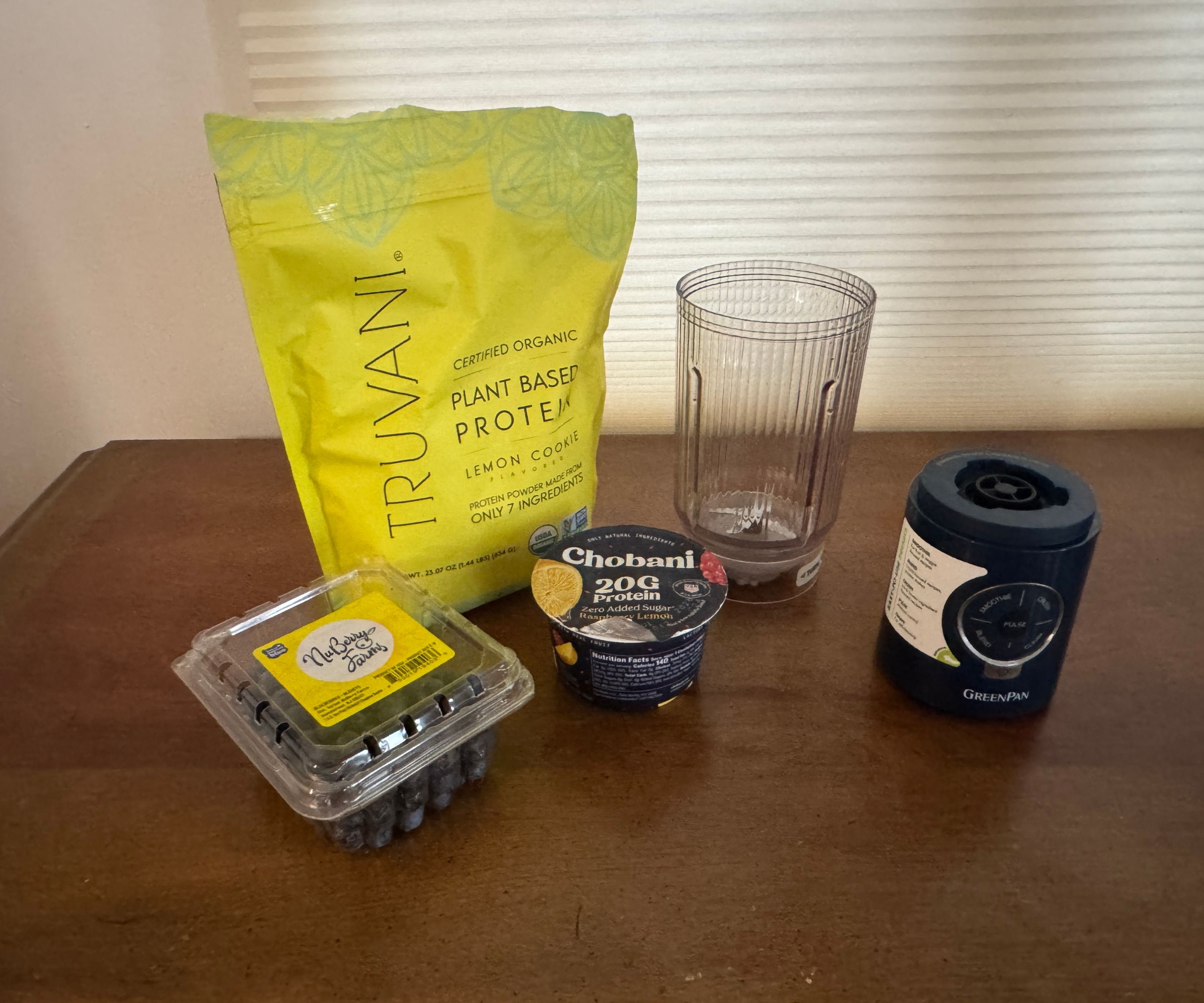 A bag of protein powder, a yogurt, and some blueberries beside a small portable blender.