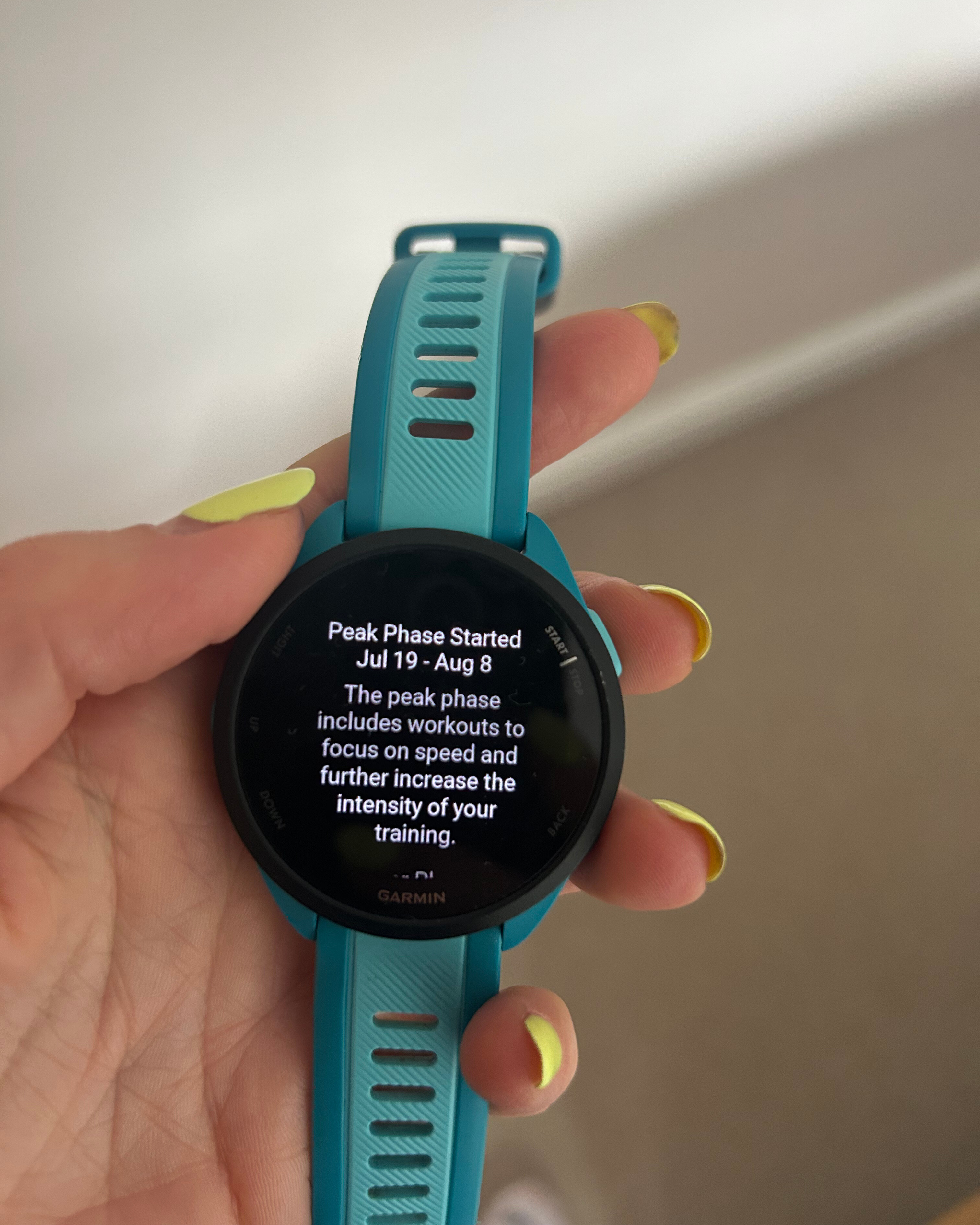 Screen of a turquoise watch held in hand. Screen reads: Peak Phase Started Jul 19 - Aug 8 The peak phase includes workouts to focus on speed and further increase the intensity of your training.