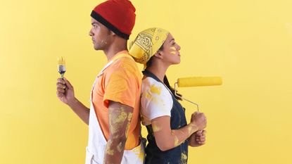 A man and woman in work clothes with paint smears all over their faces and arms stand back-to-back holding a paint brush and paint roller.
