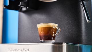 a black espresso machine by kitchenaid photographed against a blue background with silver chrome buttons and a pressure dial and steam wand