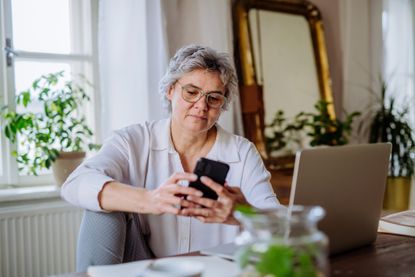 A woman in her 50s works from home. She is working in a light-filled room with plants a mirror. One leg is up on the chair and she is looking at her phone with her laptop open.