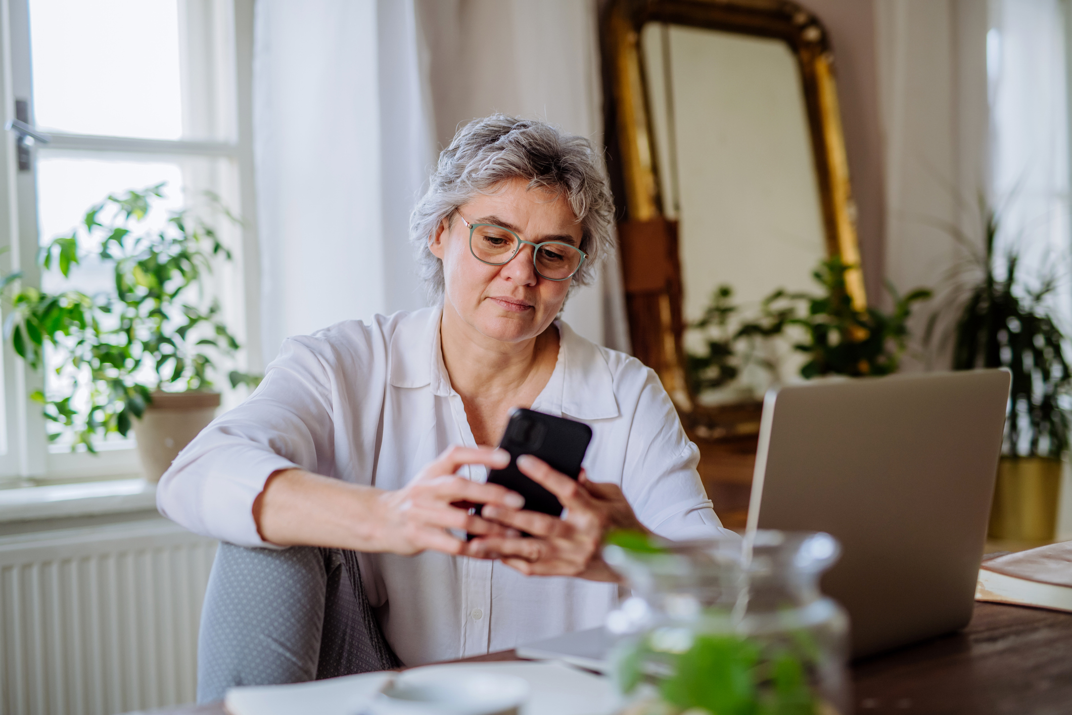 A woman in her 50s works from home. She is working in a light-filled room with plants a mirror. One leg is up on the chair and she is looking at her phone with her laptop open.