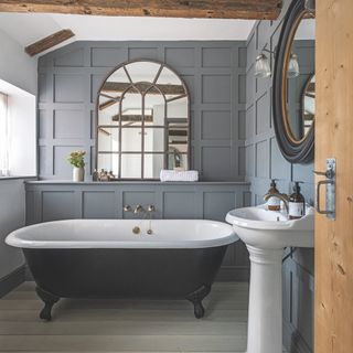 coastal bathroom with grey painted panelled walls, beams, dark grey painted freestanding bath, white painted floor, large vintage style mirror, brass taps