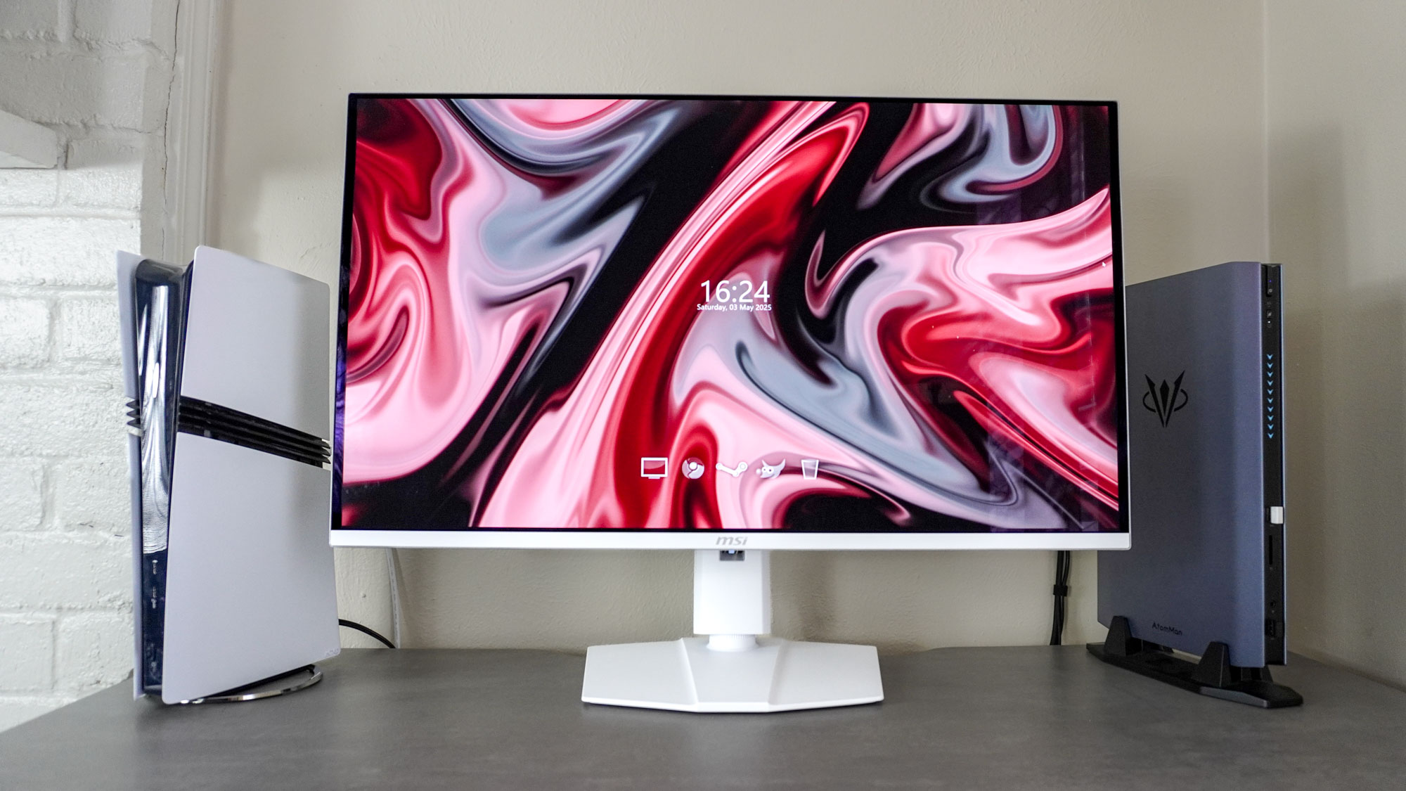 The MSI MPG 321URXW QD-OLED monitor on a desk next to a PS5 Pro and the Atomman G7 Ti mini gaming PC
